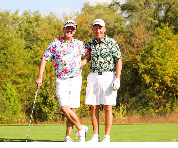 Two smiling men in colorful patterned golf shirts and white shorts stand on a green golf course with trees in the background.