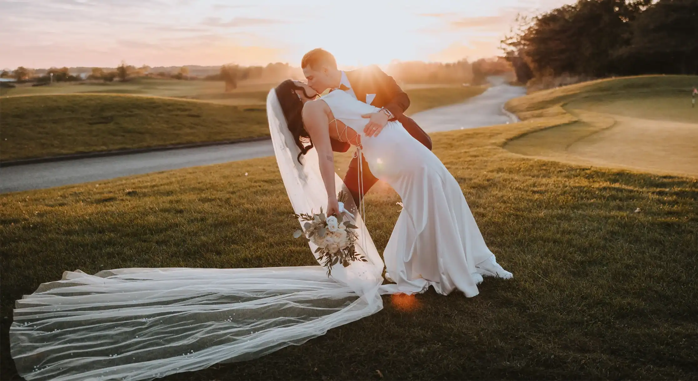 A newlywed couple kisses during sunset on a golf course. The groom dips the bride, who wears a white dress and long veil, as golden light shines behind them.