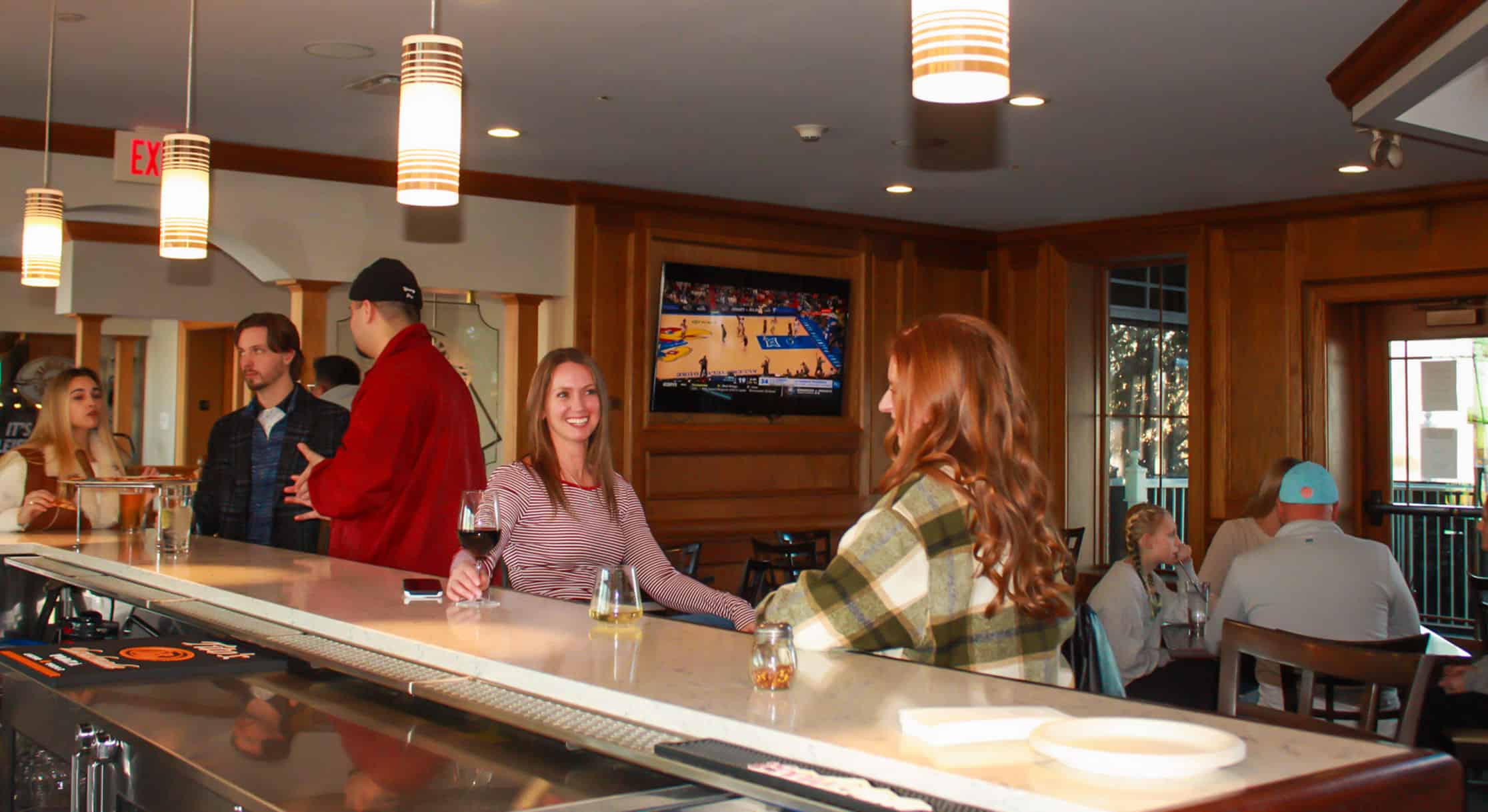 A group of people socializing at a modern bar. Two women are seated at the counter with drinks, one smiling. On the left, three people stand, one holding pizza. A TV in the background shows a basketball game.