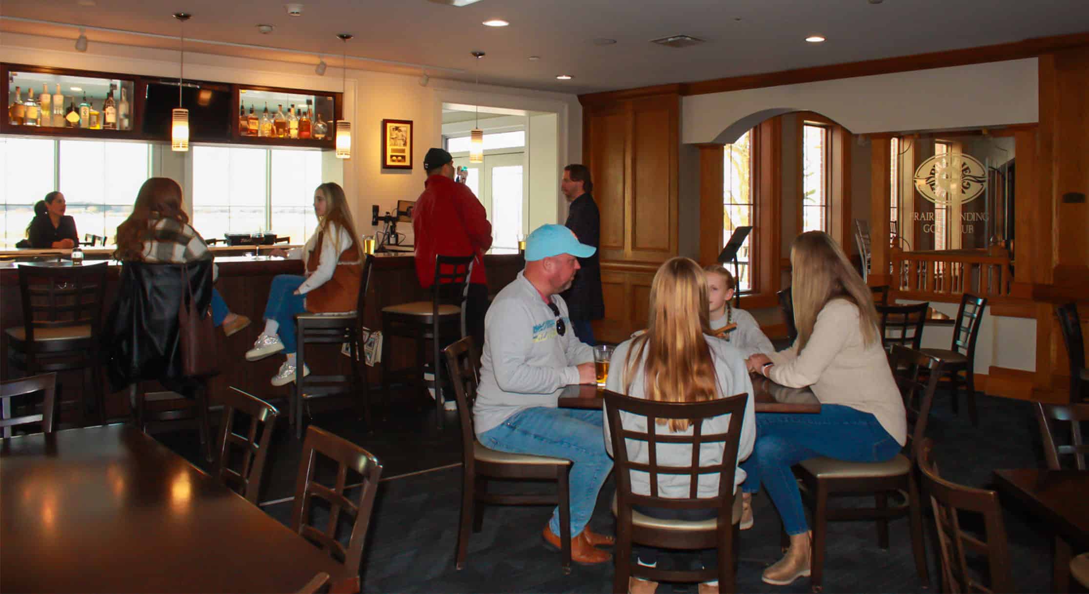 People socializing in a warmly lit bar and lounge area with wooden decor, including a well-stocked bar, tables, and chairs. A sign for 'Prairie Landing Golf Club' is visible in the background.