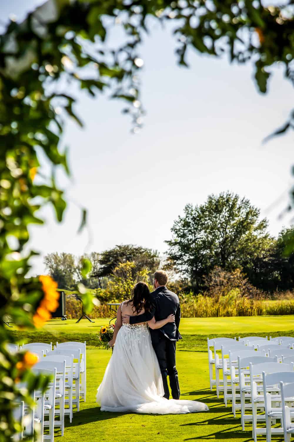 Back view of a bride in a white gown and groom in a dark suit embracing on a green lawn, framed by foliage, with rows of white chairs in the background.