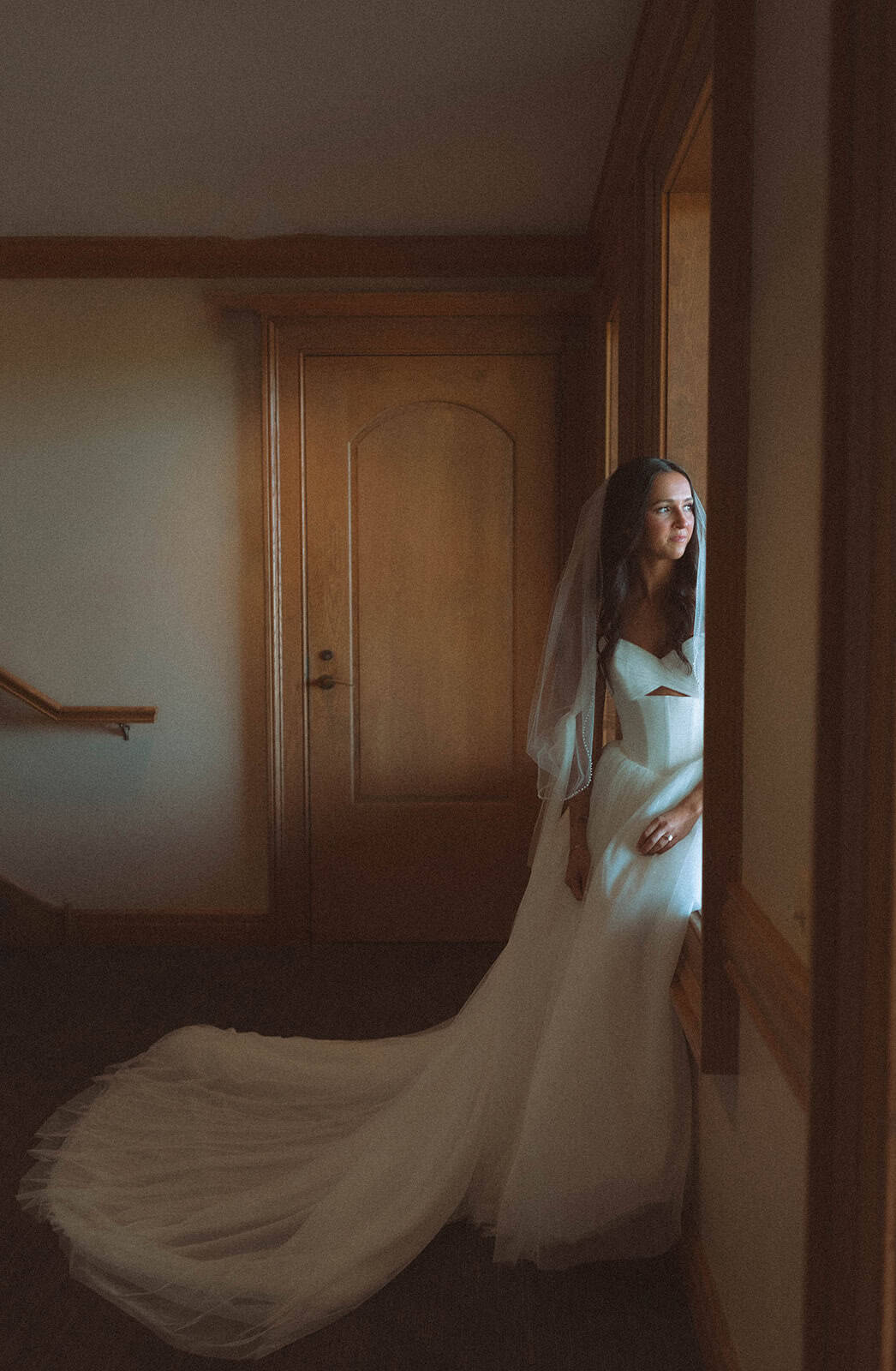 A bride in a white wedding dress and veil stands by a sunlit window, looking out thoughtfully. Her long dress train pools on the floor.