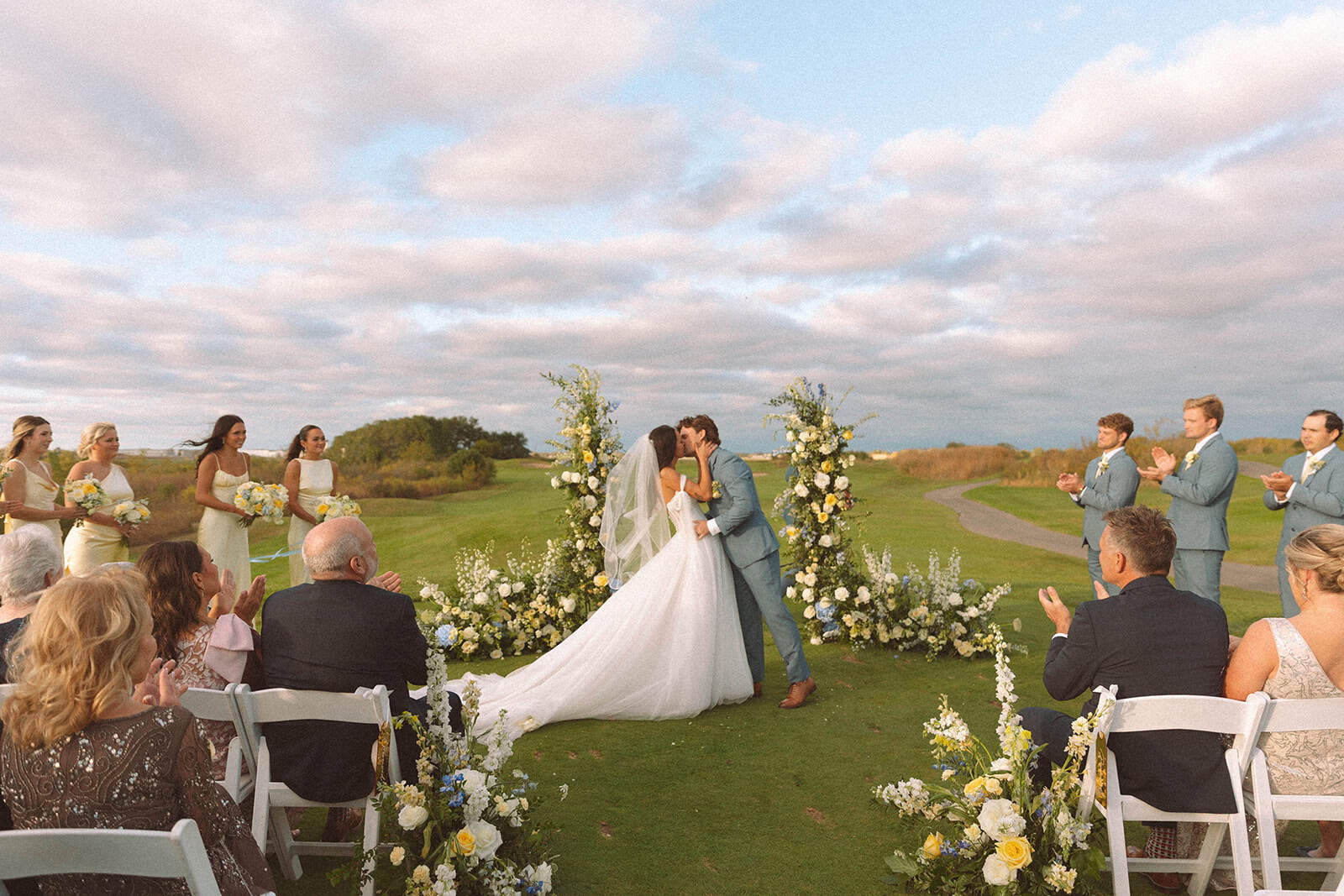 A bride and groom kiss at their outdoor wedding ceremony on a green lawn, surrounded by floral arches, clapping bridesmaids and groomsmen, and seated guests.