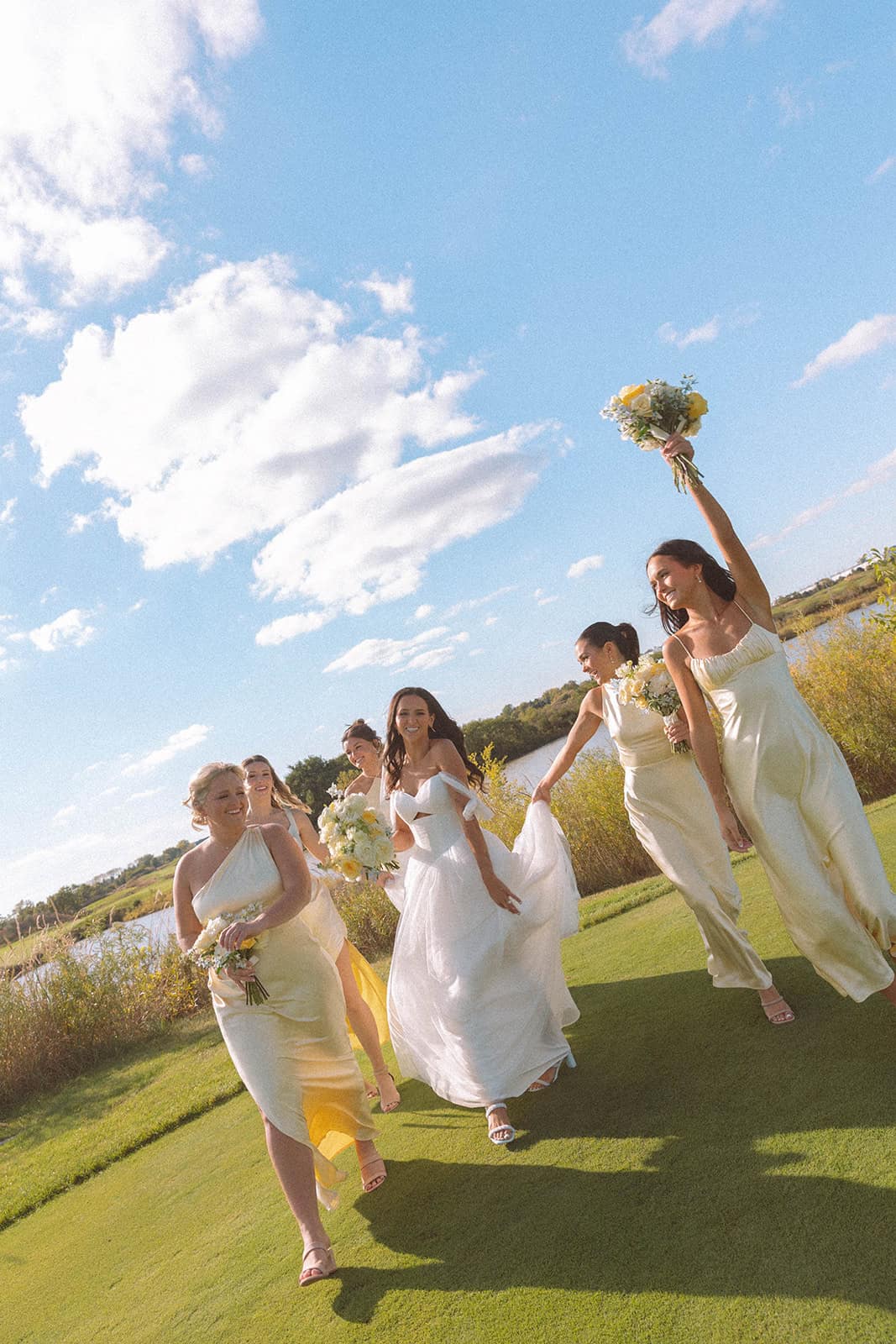 A low-angle shot of a bride in a white gown and five bridesmaids in yellow dresses walking across green grass under a blue sky, smiling and holding bouquets. One bridesmaid raises her bouquet high.