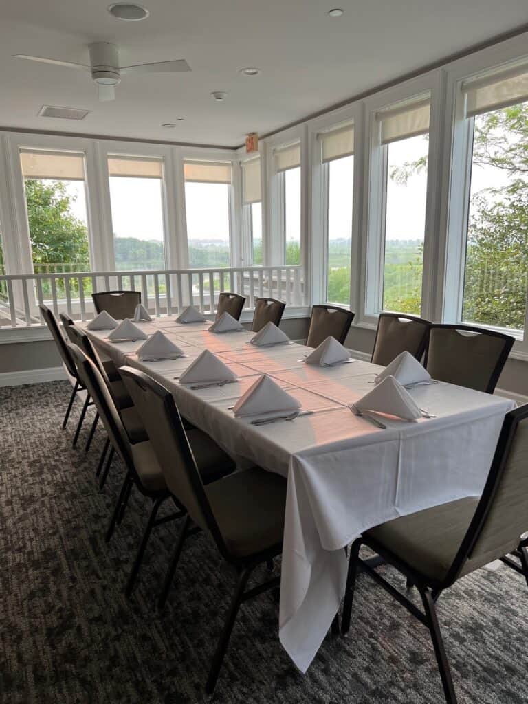 A long dining table with a white tablecloth, white napkins, and chairs, situated in a room with large windows offering a view of a green landscape.