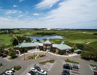 Aerial view of a large brick clubhouse with green roofs, a parking lot, a lake, and golf course fairways surrounded by green marshland under a blue sky.
