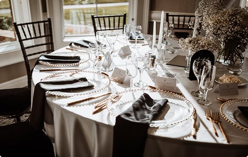 An elegantly set round dining table with a white tablecloth, clear glass charger plates with gold beaded rims, gold cutlery, dark grey napkins, white baby's breath floral centerpieces, and tall white candles, illuminated by natural light from windows.