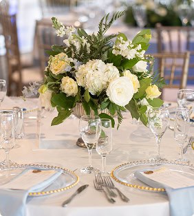 A beautifully set table with a white and yellow floral centerpiece in a silver vase, clear gold-rimmed charger plates, light blue napkins, and elegant glassware.
