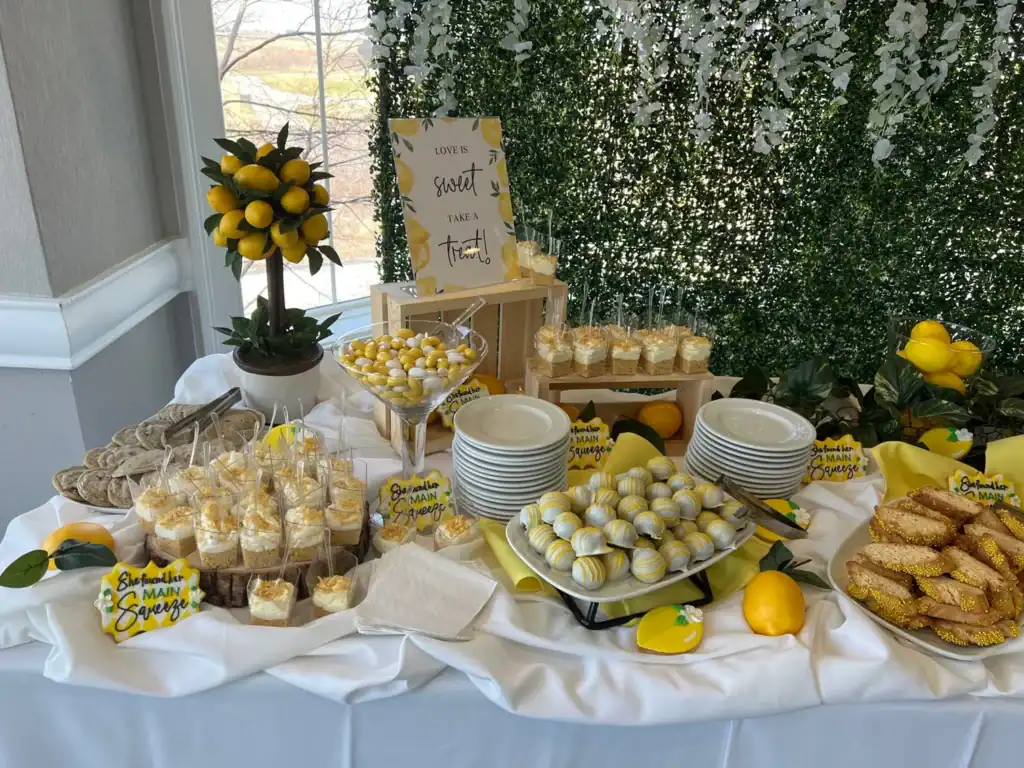 A lemon-themed dessert table featuring various yellow and white treats like layered desserts, cake pops, and cookies. Decorations include a lemon topiary, real lemons, and signs reading 'Love is sweet' and 'She found her MAIN Squeeze', all set against a green foliage wall.