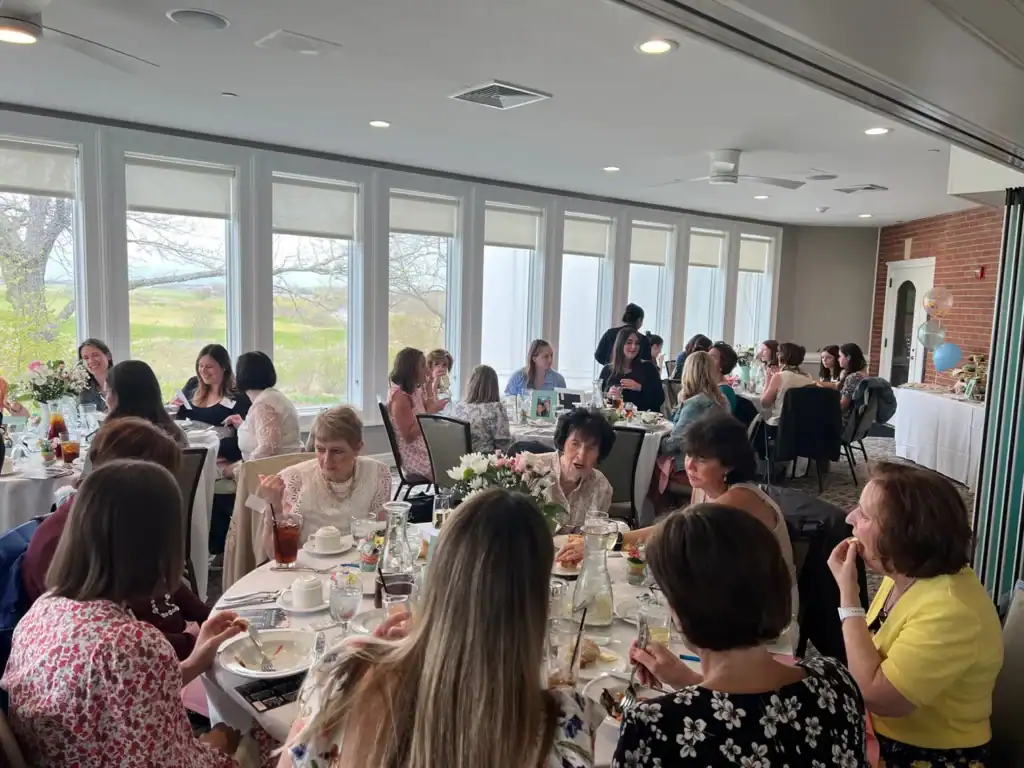 A group of women of various ages seated at round tables in a bright room with large windows overlooking a green landscape, enjoying a social luncheon.