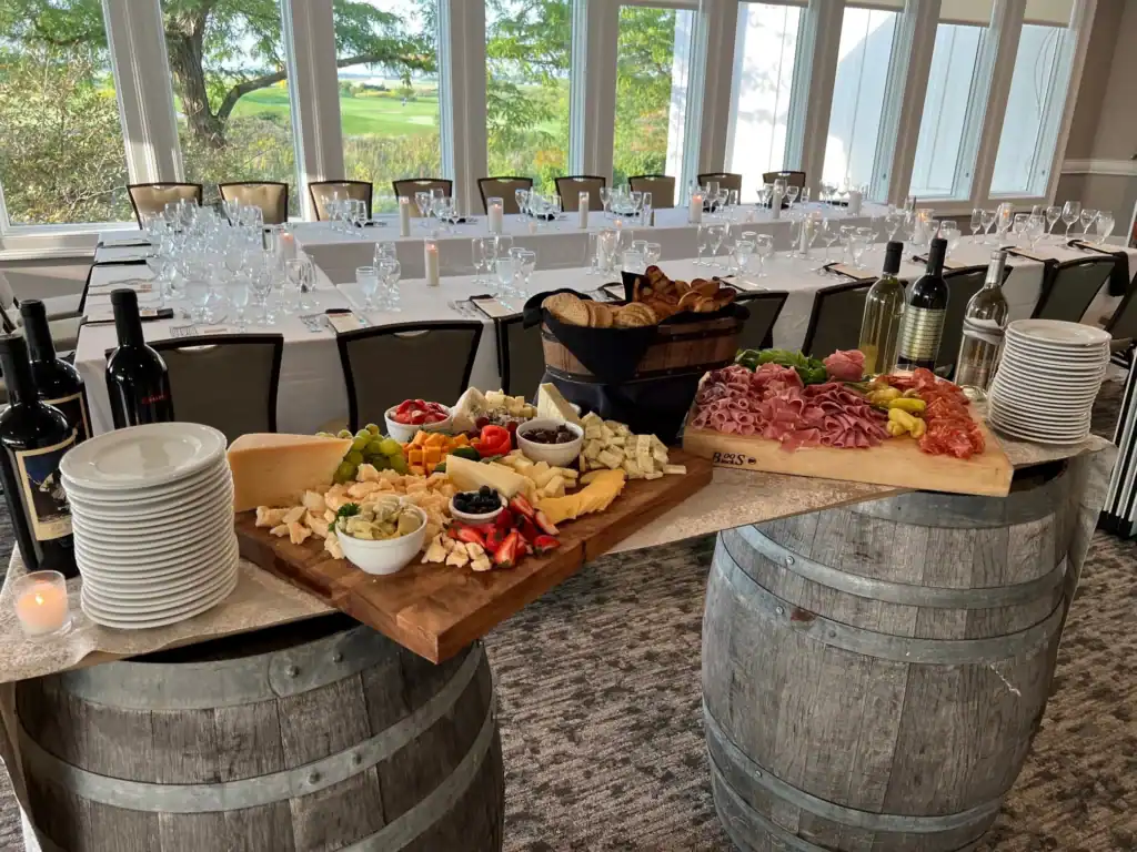 A lavish charcuterie and wine buffet set up on wooden barrels, featuring cheeses, meats, fruits, and bread. In the background, a dining table is set with glasses, overlooking a green golf course through large windows.