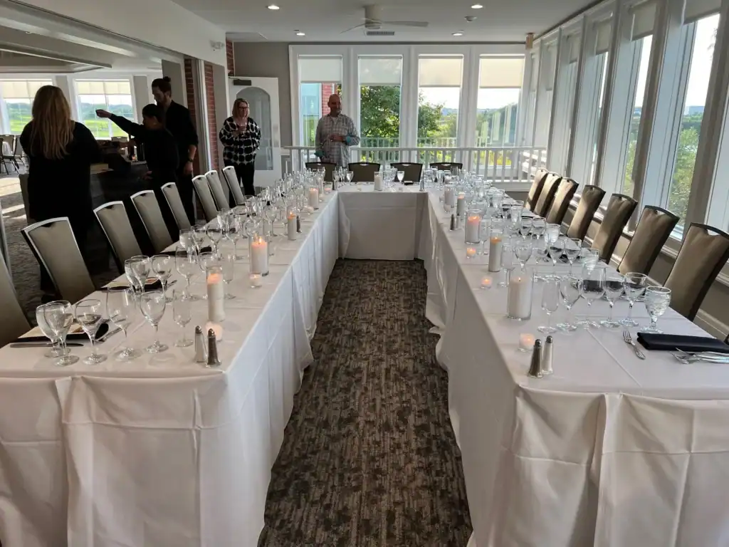 A U-shaped table set for a formal event with many wine glasses and candles, in a bright room with large windows overlooking a scenic outdoor view. Several people are visible in the background.