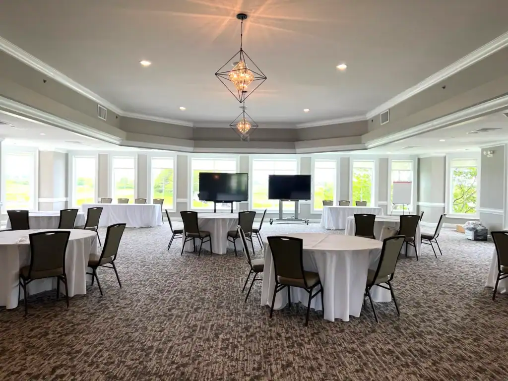 A modern event room with round tables, chairs, large windows, and two large presentation screens. Geometric chandeliers hang from the ceiling.