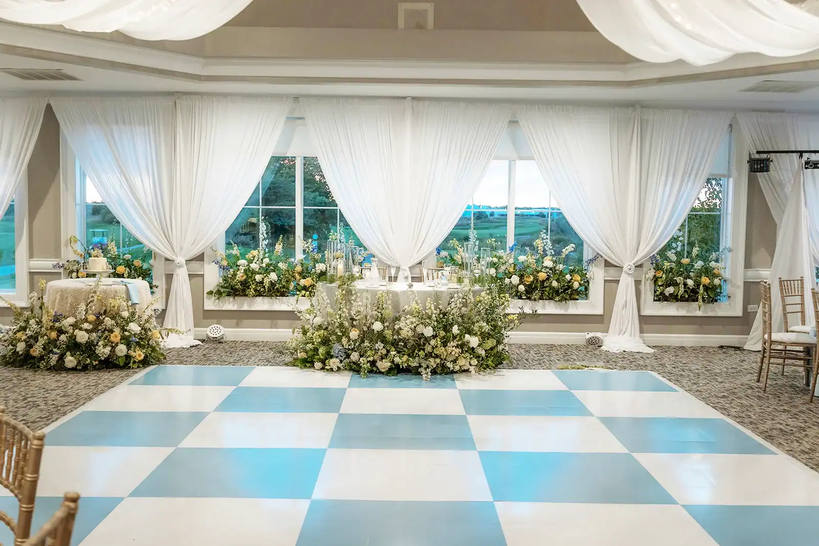 An elegant wedding reception hall with a light blue and white checkered dance floor in the foreground. The back wall features large windows draped with white sheer curtains, framed by lush floral arrangements in white, cream, yellow, and blue. A sweetheart table with cascading flowers is centered against the windows, and a cake table with a white wedding cake is on the left, also surrounded by flowers.