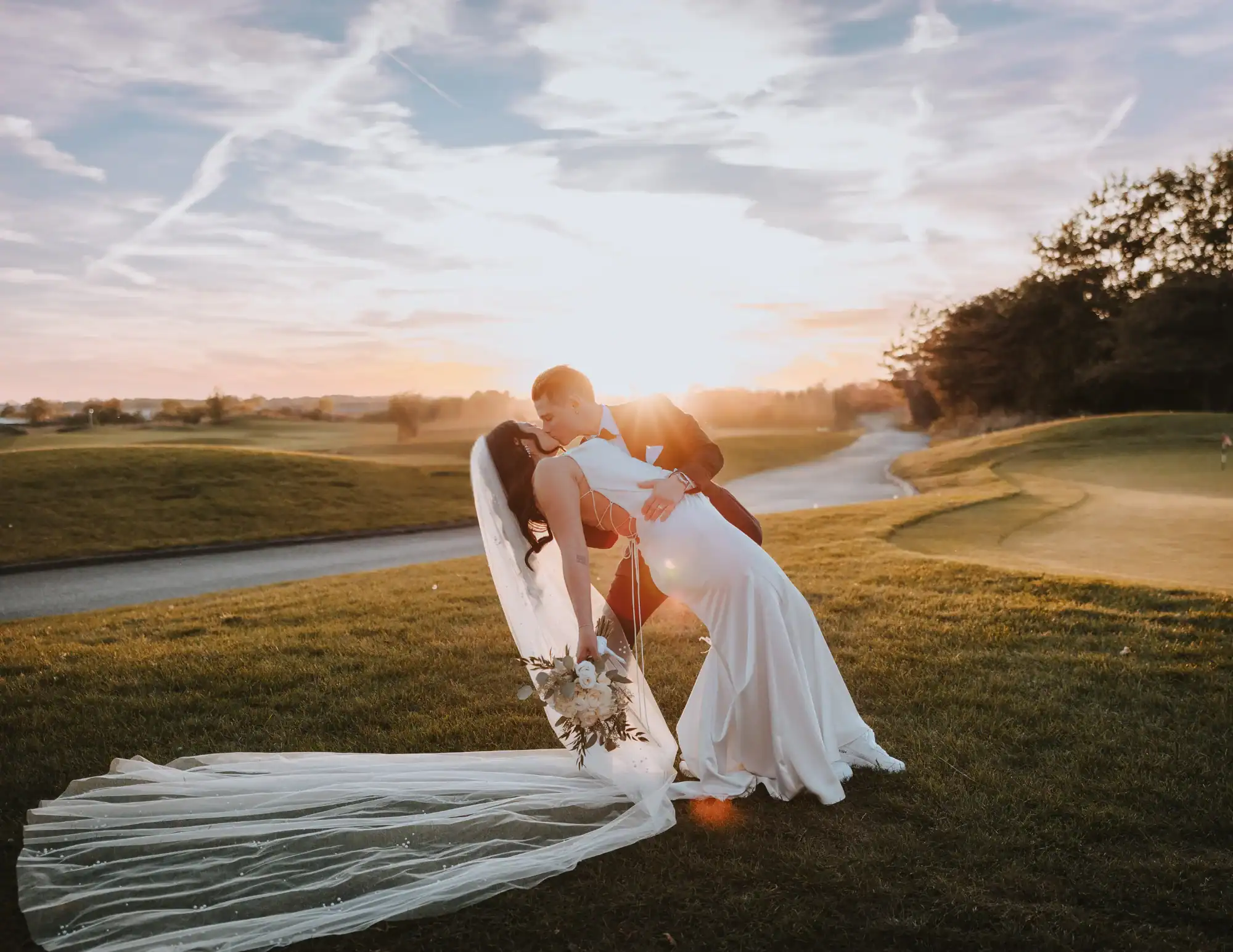 A newlywed couple kissing during sunset on a golf course. The groom dips the bride, who wears a white dress and long veil, as the sun creates a warm glow behind them.
