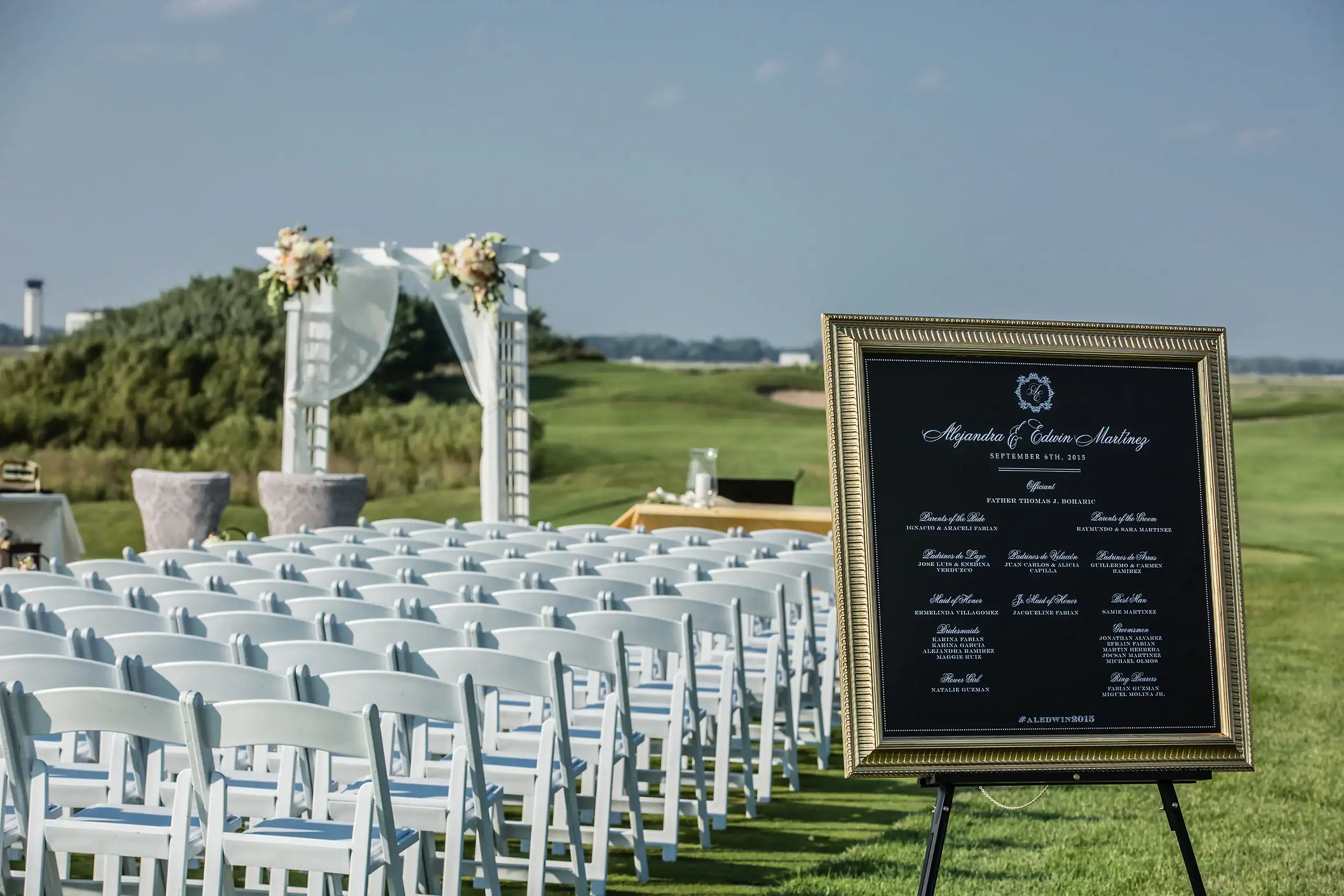 An outdoor wedding ceremony setup on a green lawn, featuring rows of white chairs, a white floral arch, and a black and gold framed sign detailing the wedding party for Alejandra & Edwin Martinez.