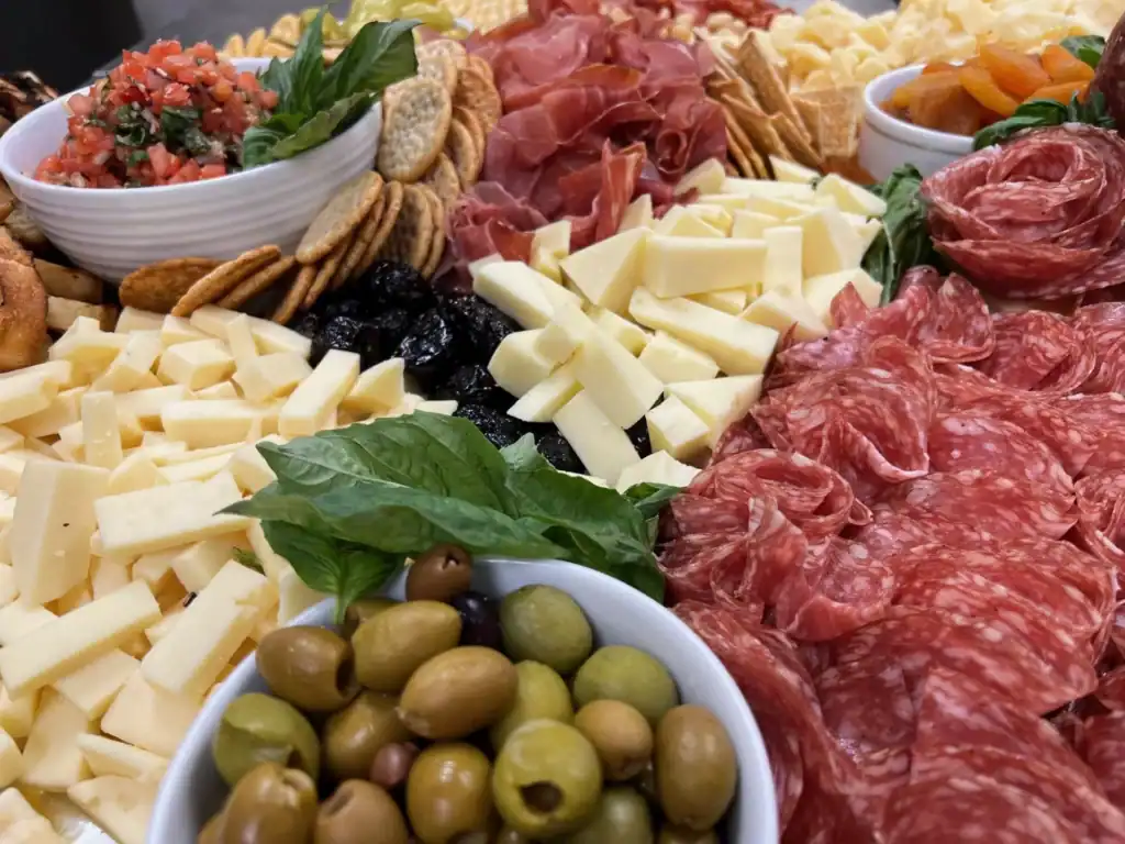 A large, abundant charcuterie board filled with various cheeses, cured meats, crackers, olives, bruschetta, dried apricots, and dried plums, garnished with fresh basil.