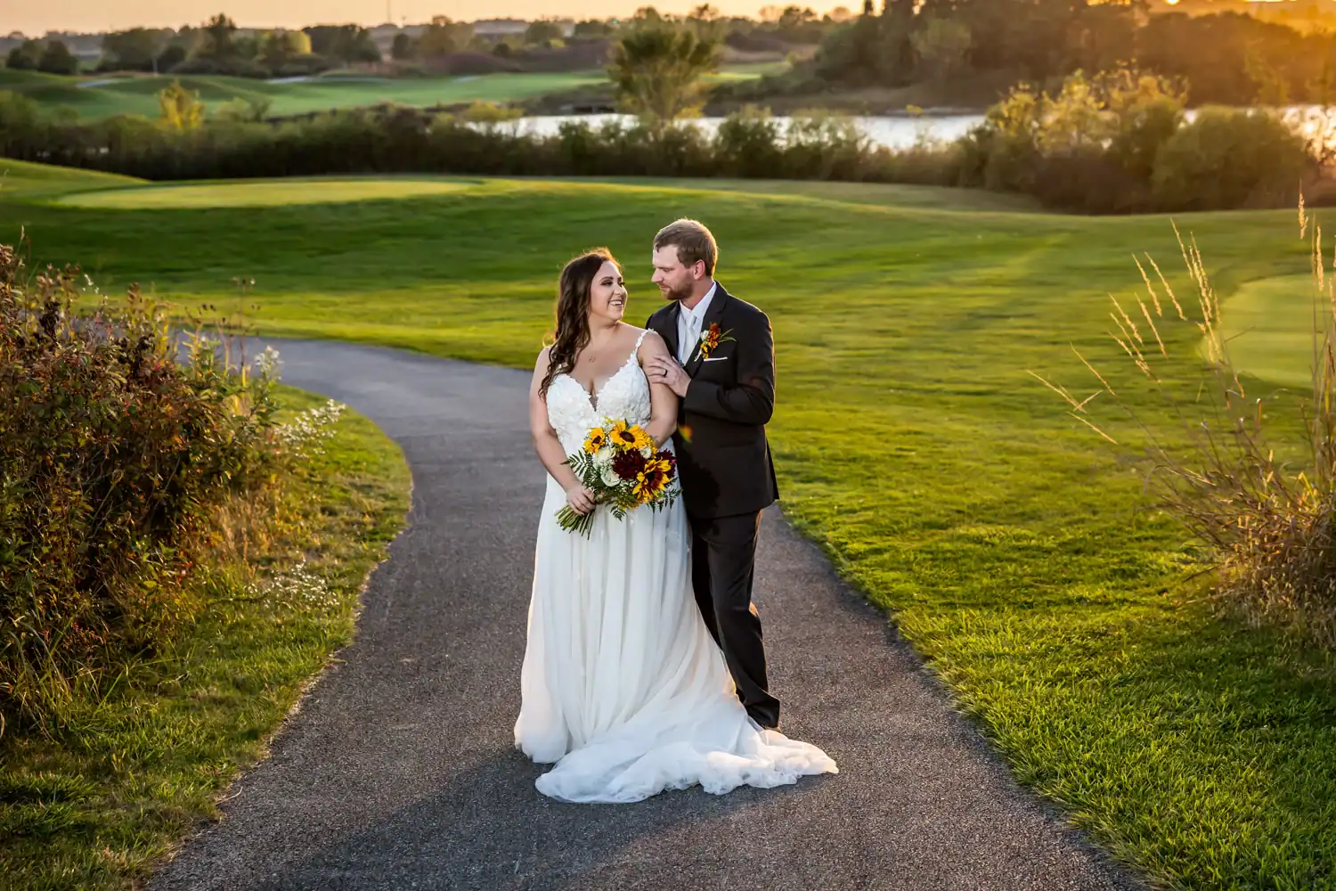 A newlywed couple, a bride in a white gown holding sunflowers and a groom in a dark suit, stand on a paved path, gazing at each other and smiling. They are in a lush green outdoor setting with a lake and trees in the background, bathed in warm golden hour light.