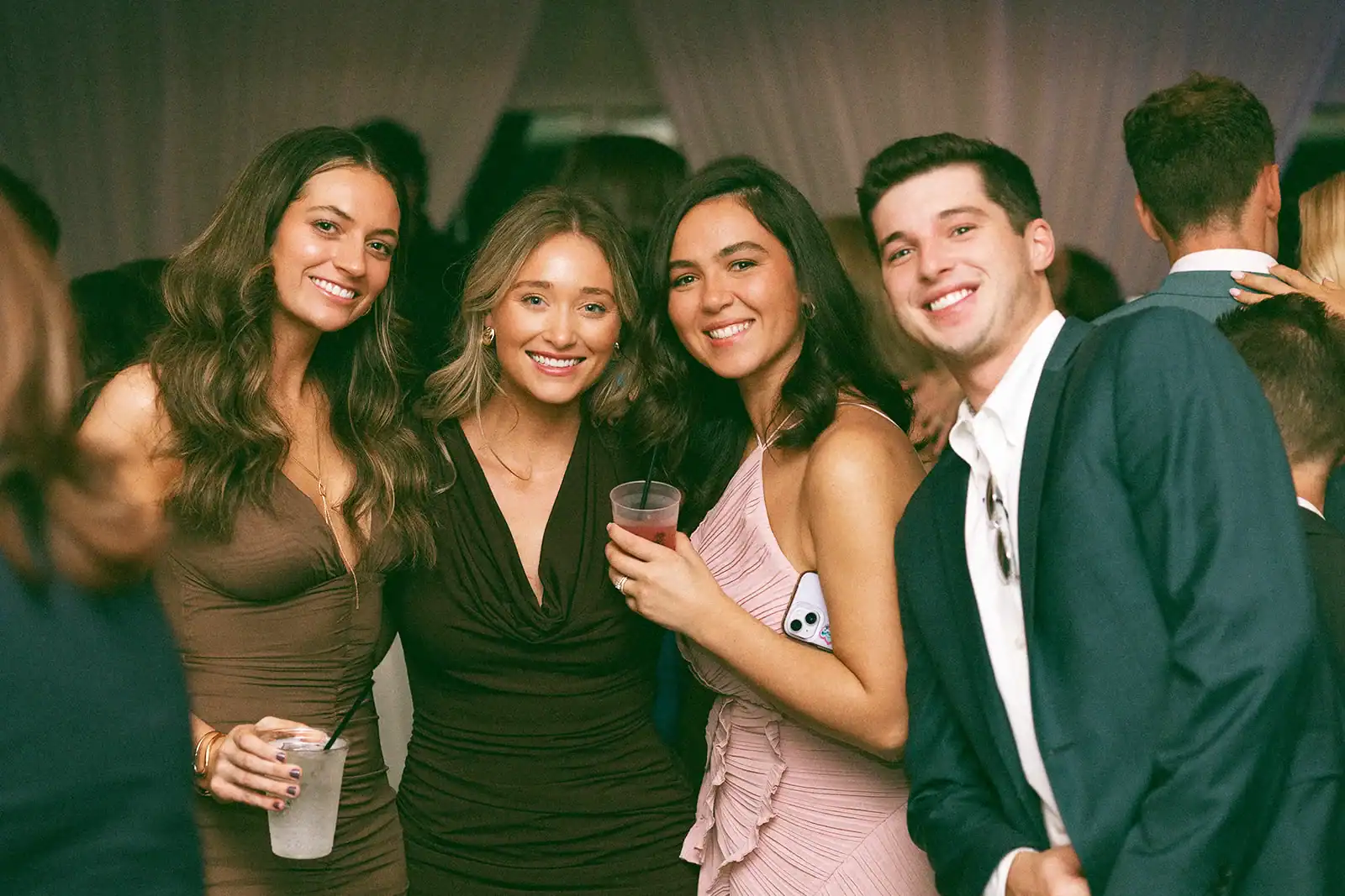 Four young adults, three women and one man, smiling brightly at the camera at a party. They are dressed in semi-formal attire and appear to be enjoying themselves.
