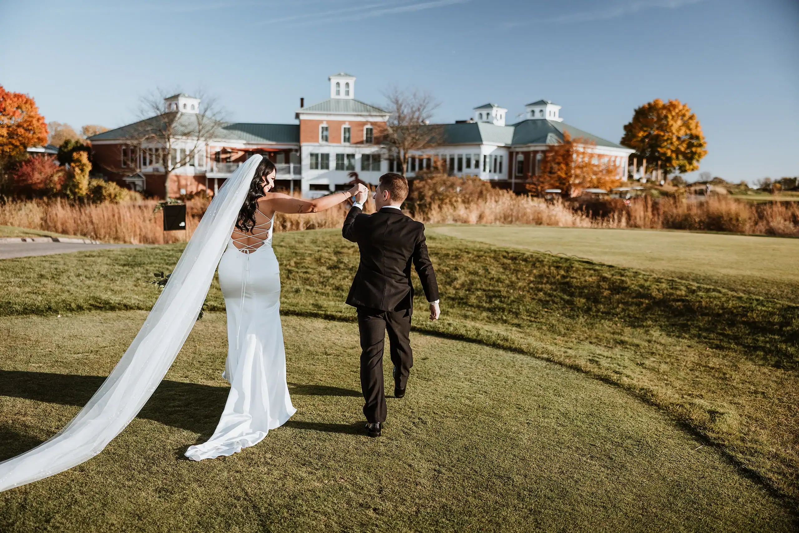 A newlywed couple, seen from behind, walking hand-in-hand across a green lawn towards a grand building with autumn trees. The bride wears a white gown and long veil, the groom a black suit.