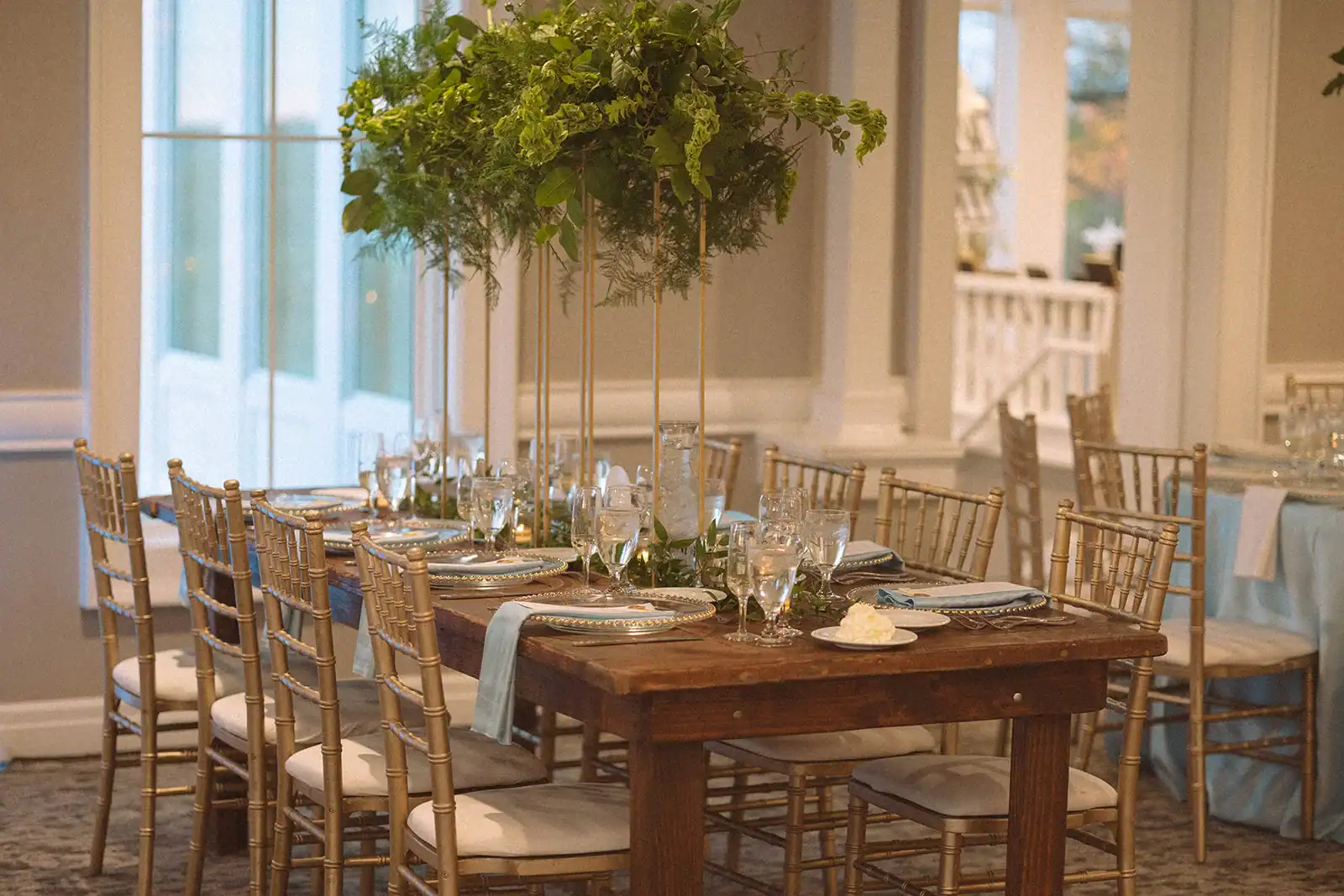 A long wooden dining table set for a formal event, featuring tall gold centerpieces with lush green foliage, gold Chiavari chairs, and place settings with clear glass chargers, white plates, and light blue napkins. Windows are visible in the background.