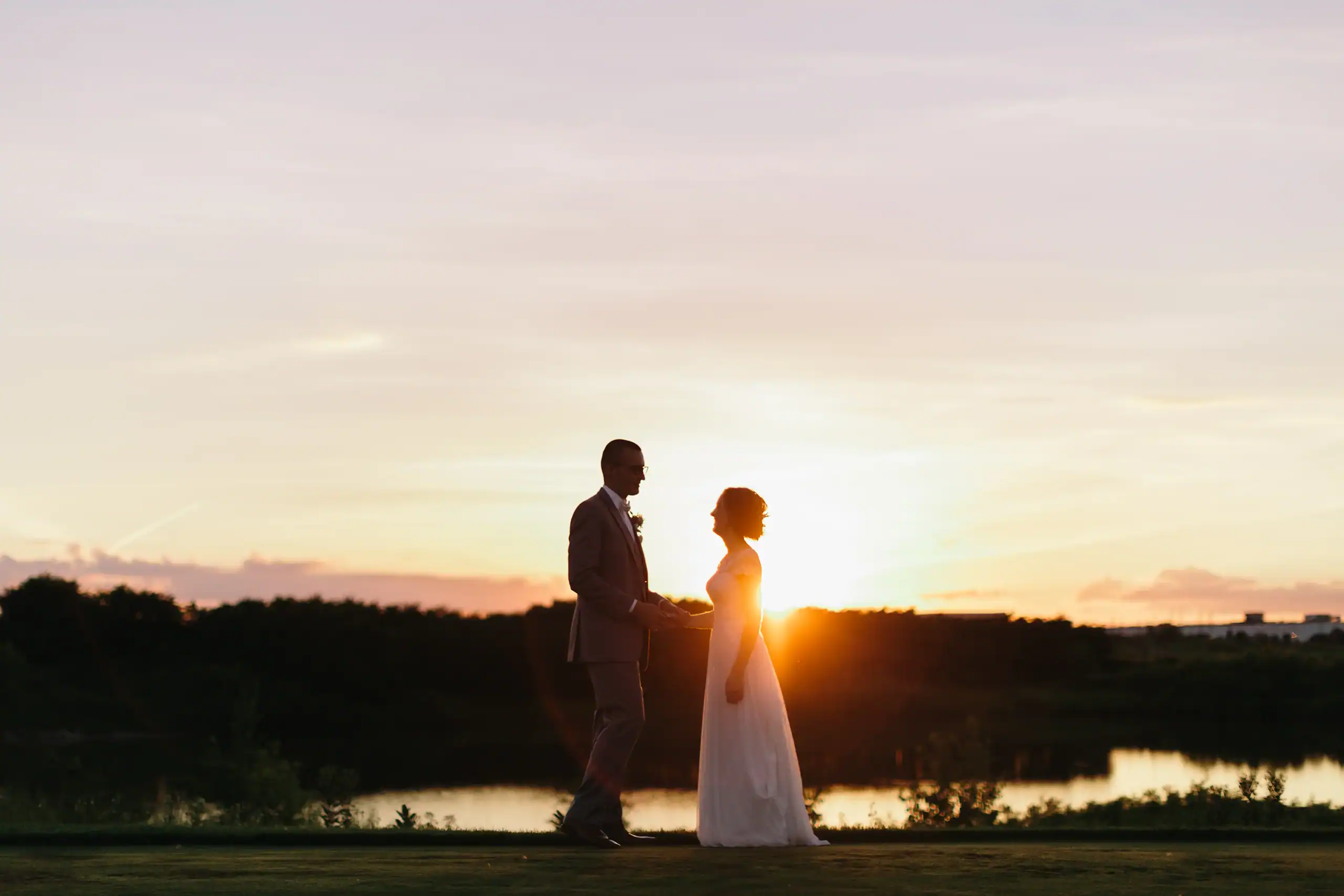 Silhouette of a bride and groom holding hands and looking at each other against a vibrant sunset over a lake.