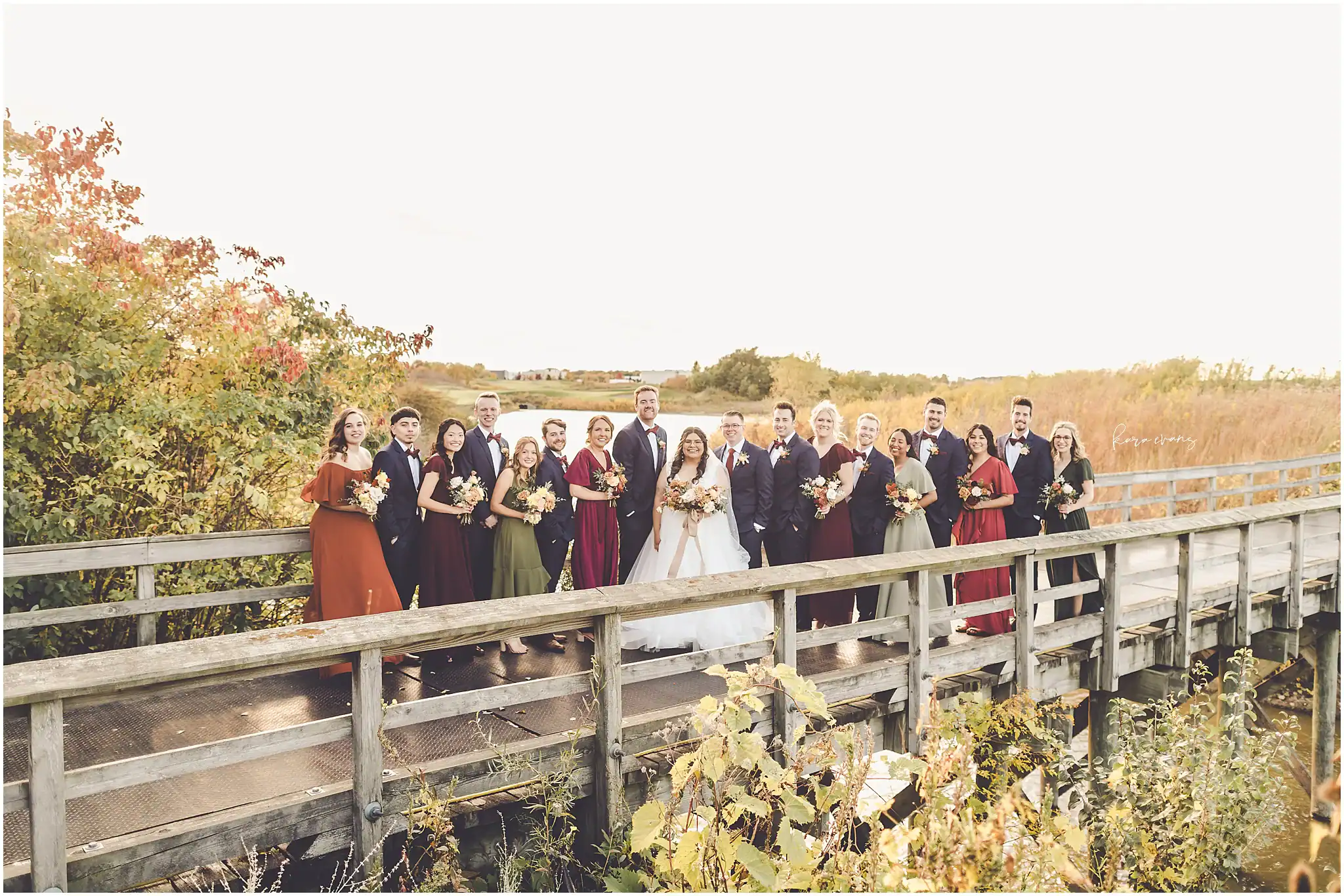 A large wedding party, including the bride in a white gown and groom in a dark suit, stands smiling on a wooden bridge. Bridesmaids wear dresses in burnt orange, burgundy, and olive green, while groomsmen wear dark suits with burgundy bow ties. The background features vibrant autumn foliage and golden marsh grasses.