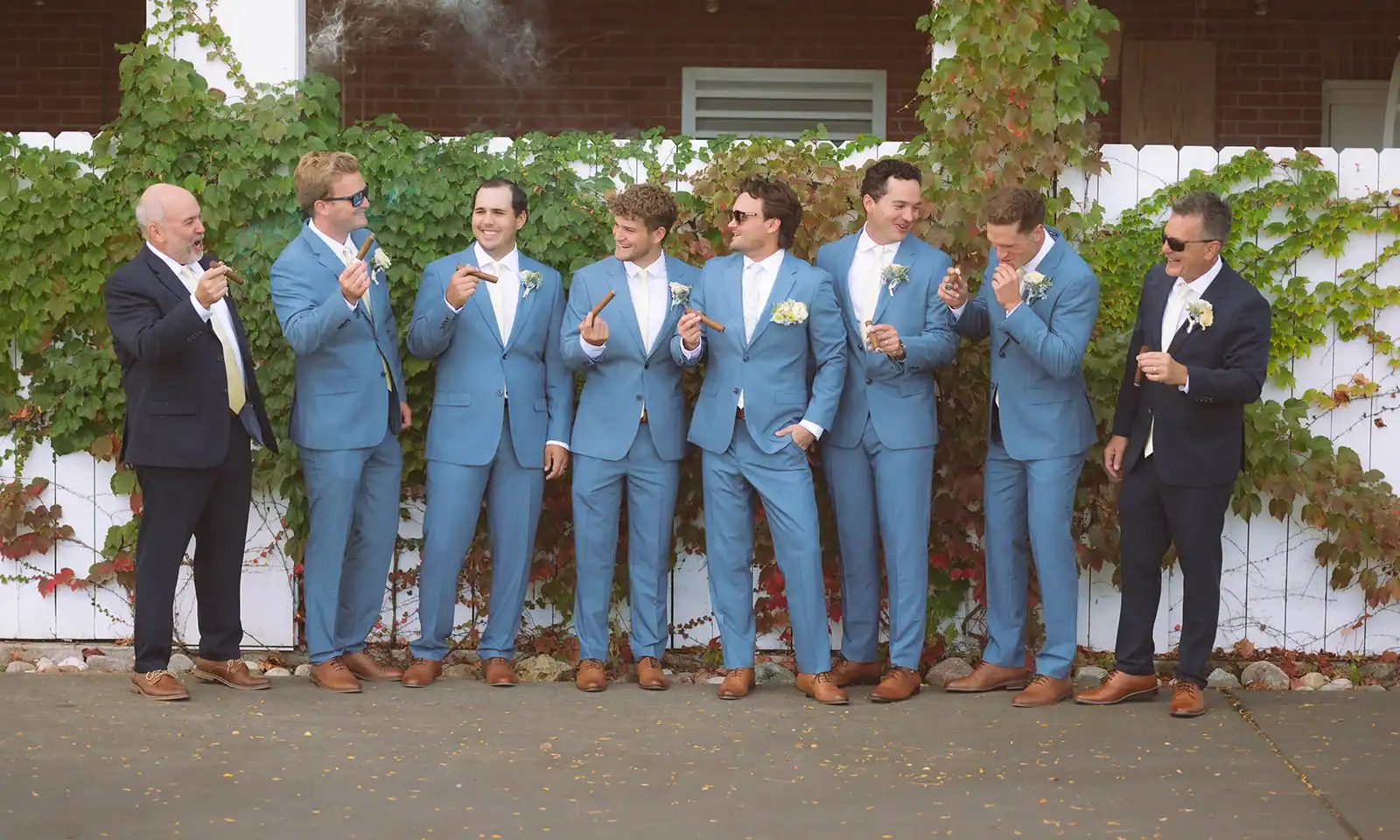 Eight men, including a groom and groomsmen in blue suits and two older men in dark suits, standing and laughing while smoking cigars in front of an ivy-covered white fence.