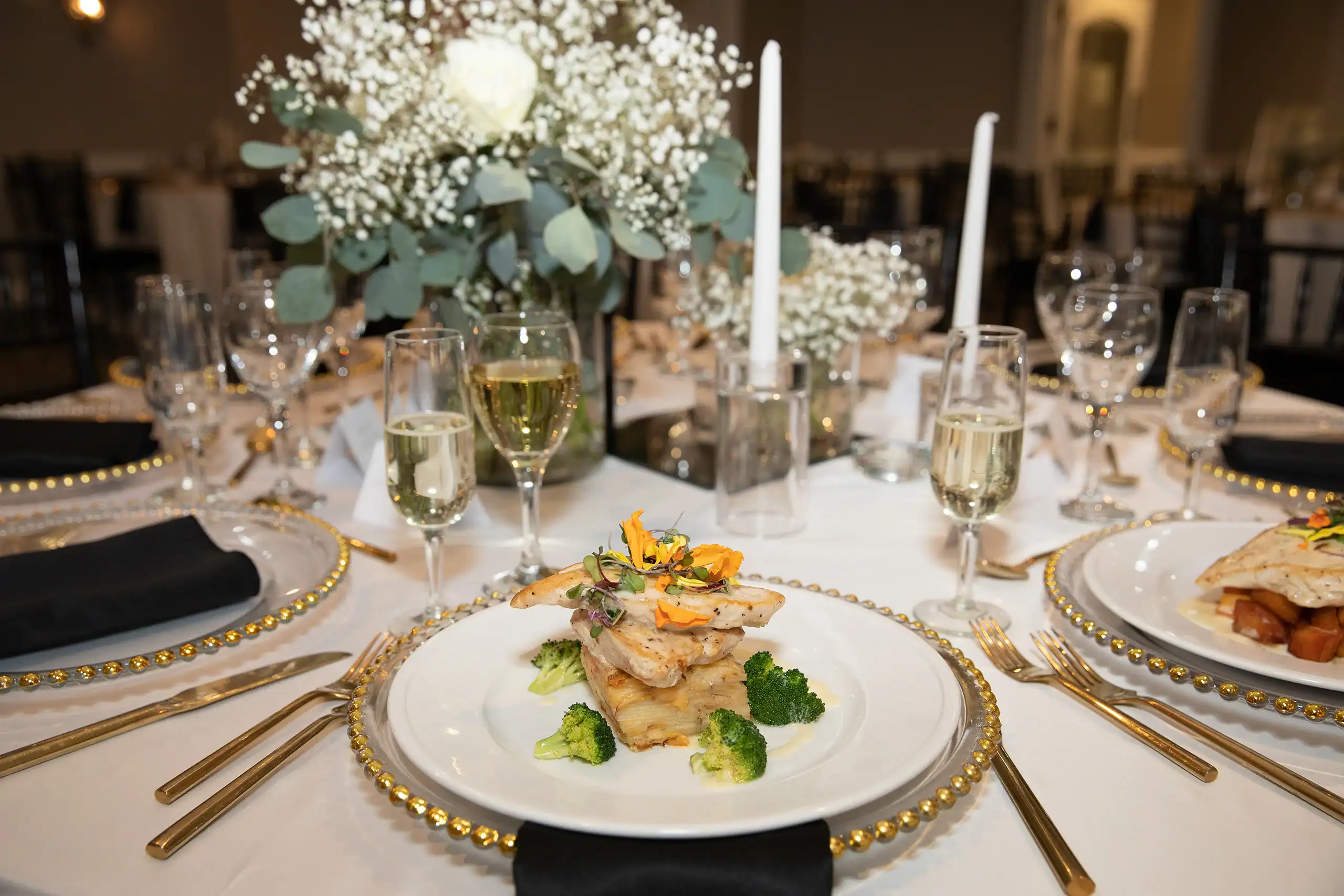A formal dining table set with a gourmet meal of chicken or fish, potato gratin, and broccoli, alongside champagne glasses, gold cutlery, and a floral centerpiece with white roses and baby's breath.