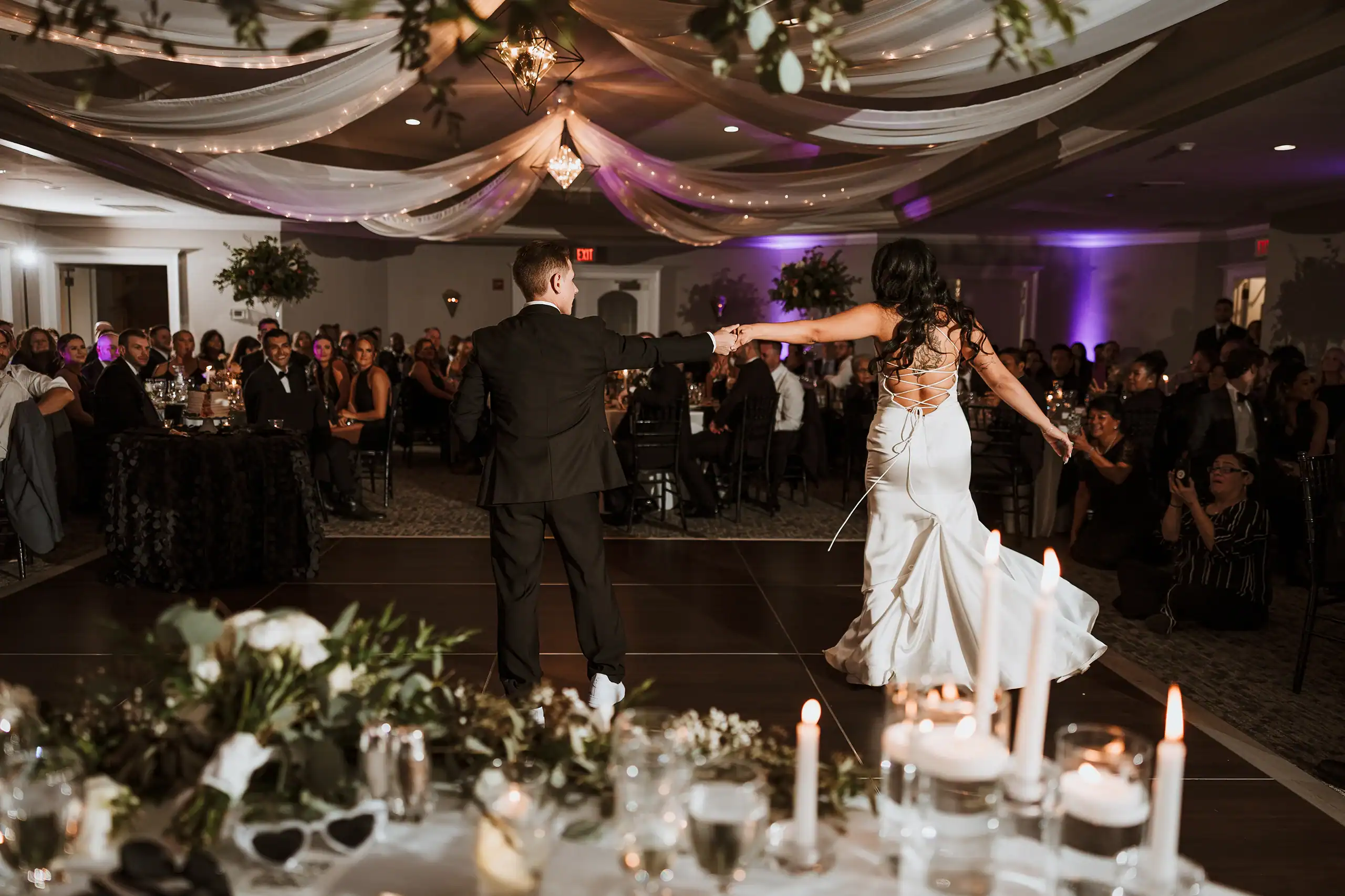A couple in formal wear, seen from behind, holding hands and dancing on a dark dance floor at a wedding reception. The male wears a black suit, the female a white backless gown. Guests are seated around them, and the ceiling is draped with fabric and string lights.