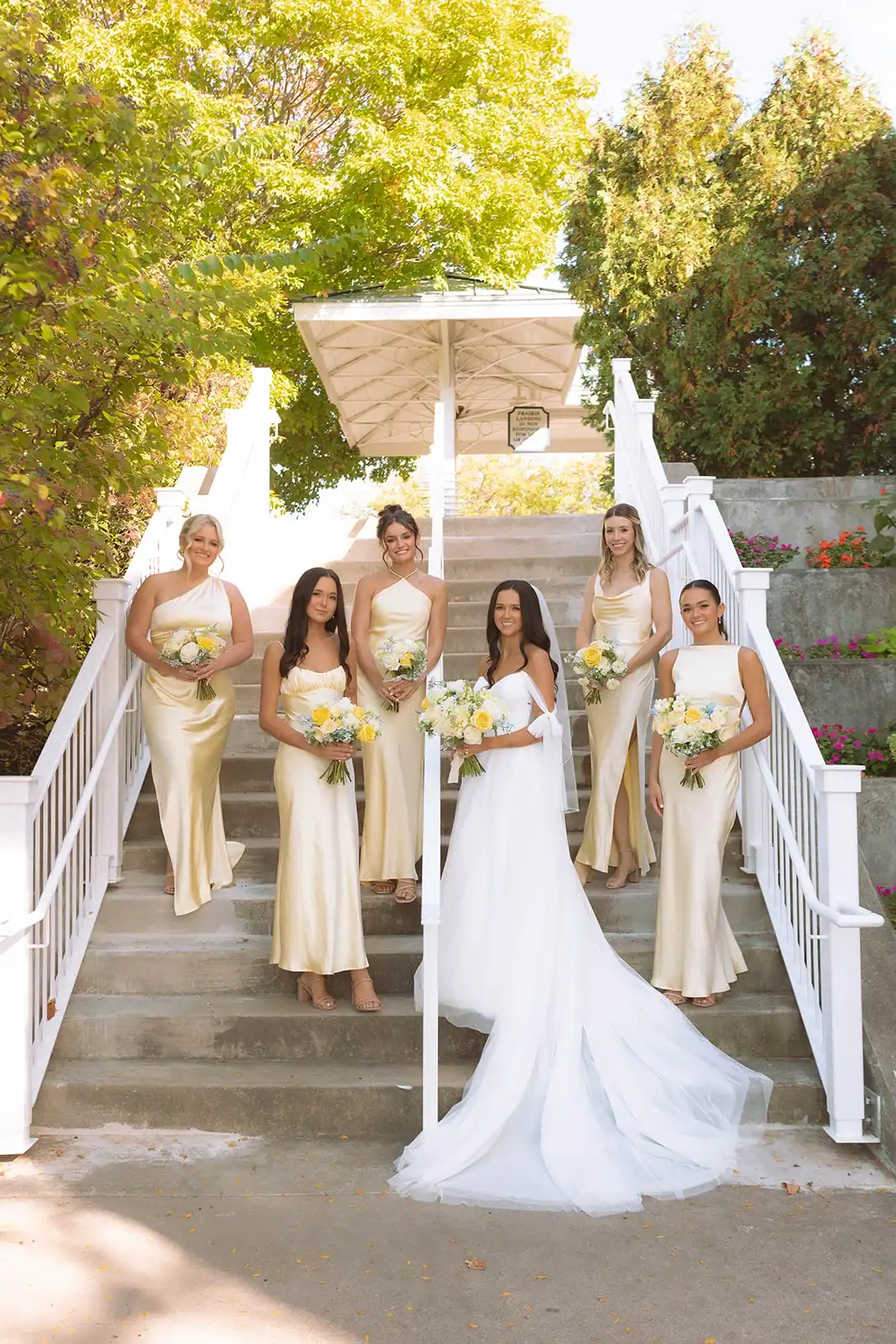 A bride in a white gown and five bridesmaids in light yellow dresses pose on a concrete staircase with white railings, surrounded by green trees and a white gazebo.