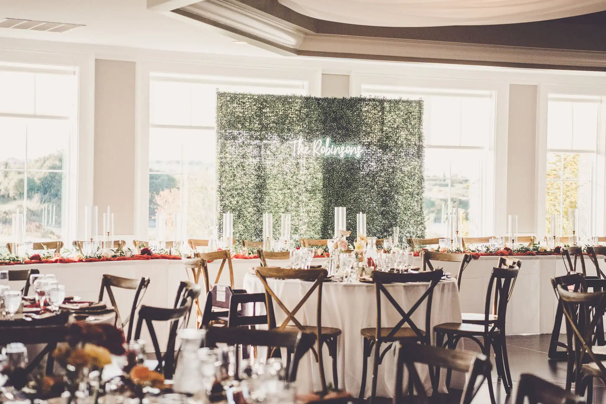 An elegant wedding reception room with a green hedge wall featuring a 'The Robinsons' neon sign, white tables with floral centerpieces, and wooden cross-back chairs.