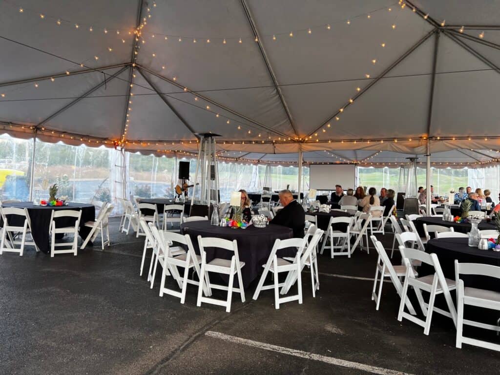Interior of a large event tent with string lights, round tables with black tablecloths, white folding chairs, and guests attending a gathering.