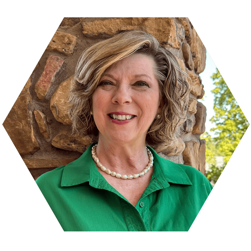 A portrait of a Teresa Ham, wearing a green blouse and pearl necklace, standing in front of a stone wall with trees in the background.