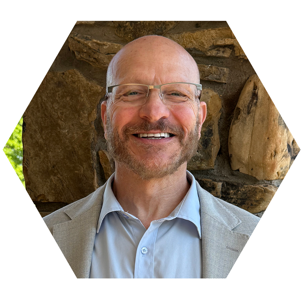 A portrait of a smiling middle-aged man with glasses, wearing a light-colored blazer and shirt, standing in front of a rustic stone wall background.
