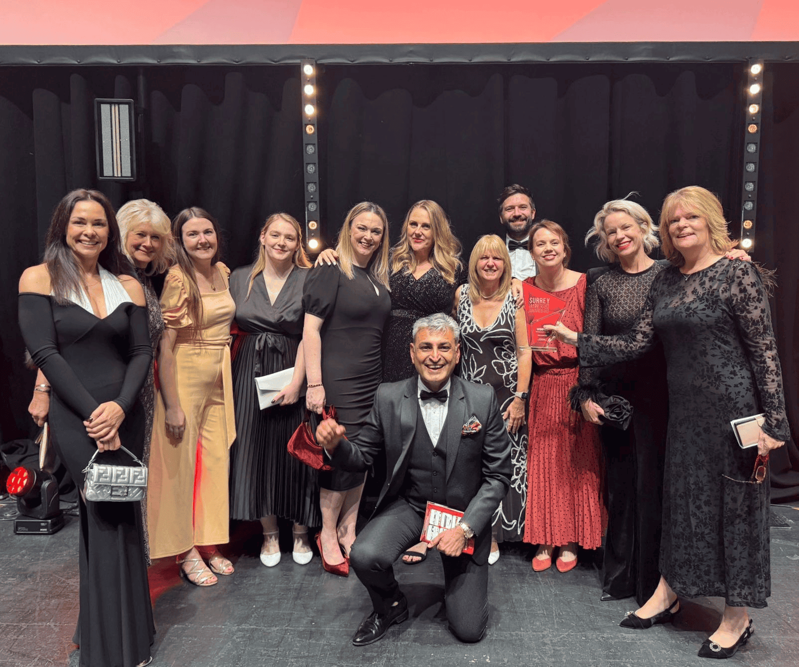 A group of professionally dressed people at a formal event, smiling on stage with a black backdrop and stage lights, showcasing a celebratory occasion.