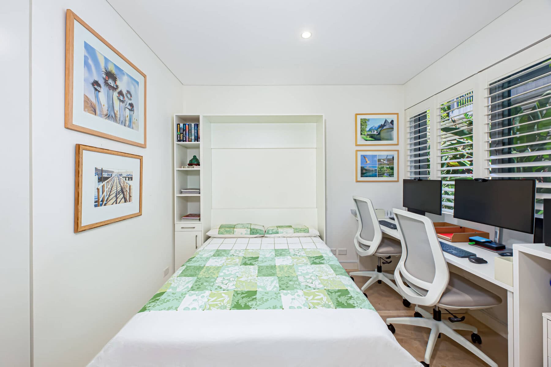 A bright, tidy bedroom with a green-and-white patchwork bedspread, built-in shelves, framed wall art, and a double desk with two office chairs and dual monitors by a window with white shutters.