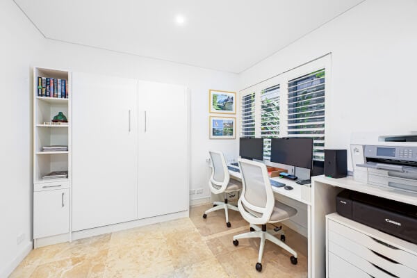 A bright, modern home office with two white desks, dual monitors, two white chairs, a printer, and a cabinet. Shelves hold books and decor; windows with shutters let in natural light.