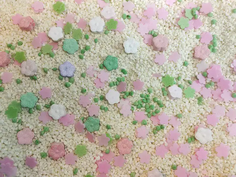 A visually stunning mix of custom cat litter deodorizer sprinkles, including pink flower flakes, candy shapes, and green granules, scattered on a bed of natural plant-based cassava cat litter. This showcases the beautiful customization options a manufacturer offers to enhance odor control and create a signature product.