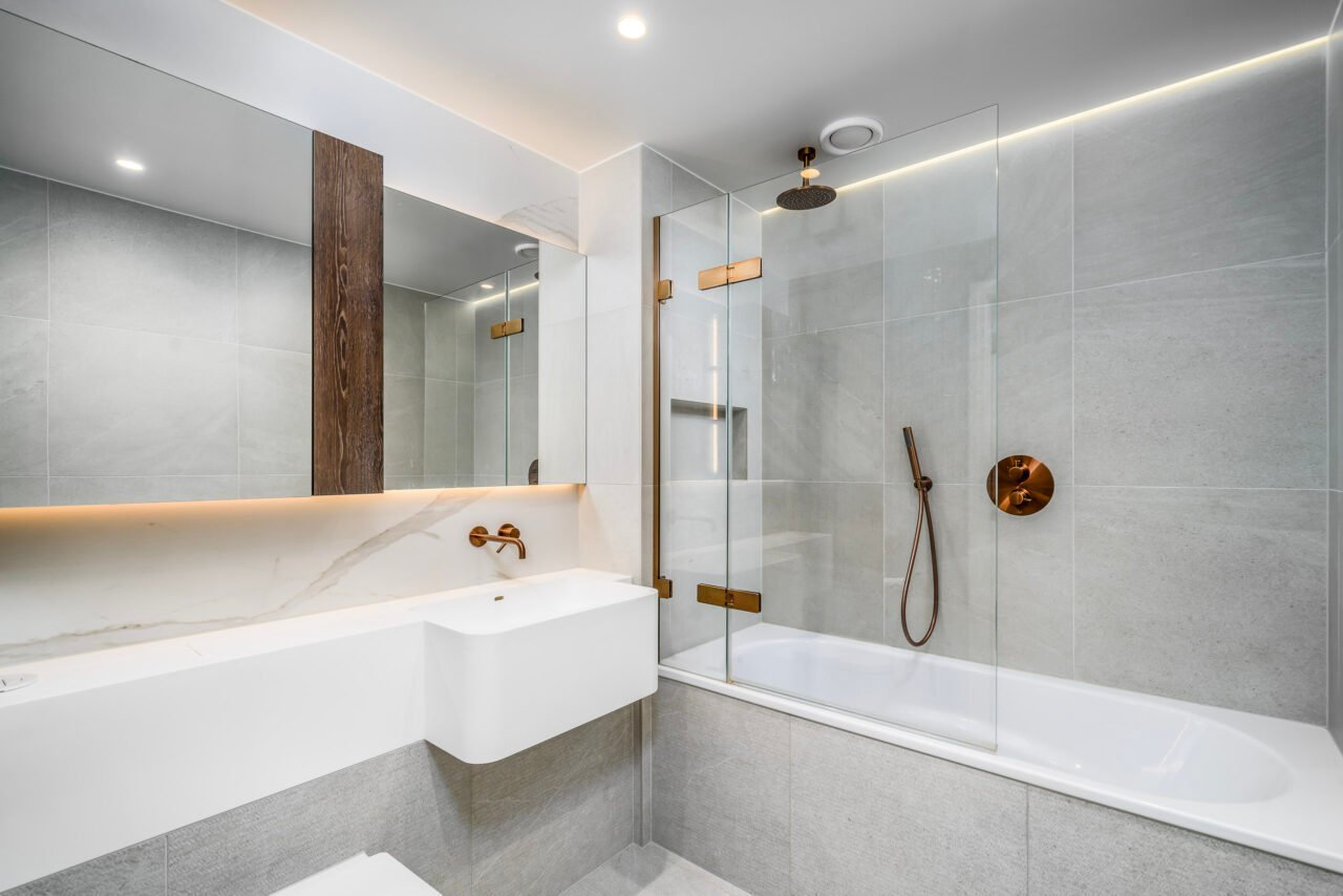 A modern grey tiled bathroom with a glass shower enclosure and white bathtub.