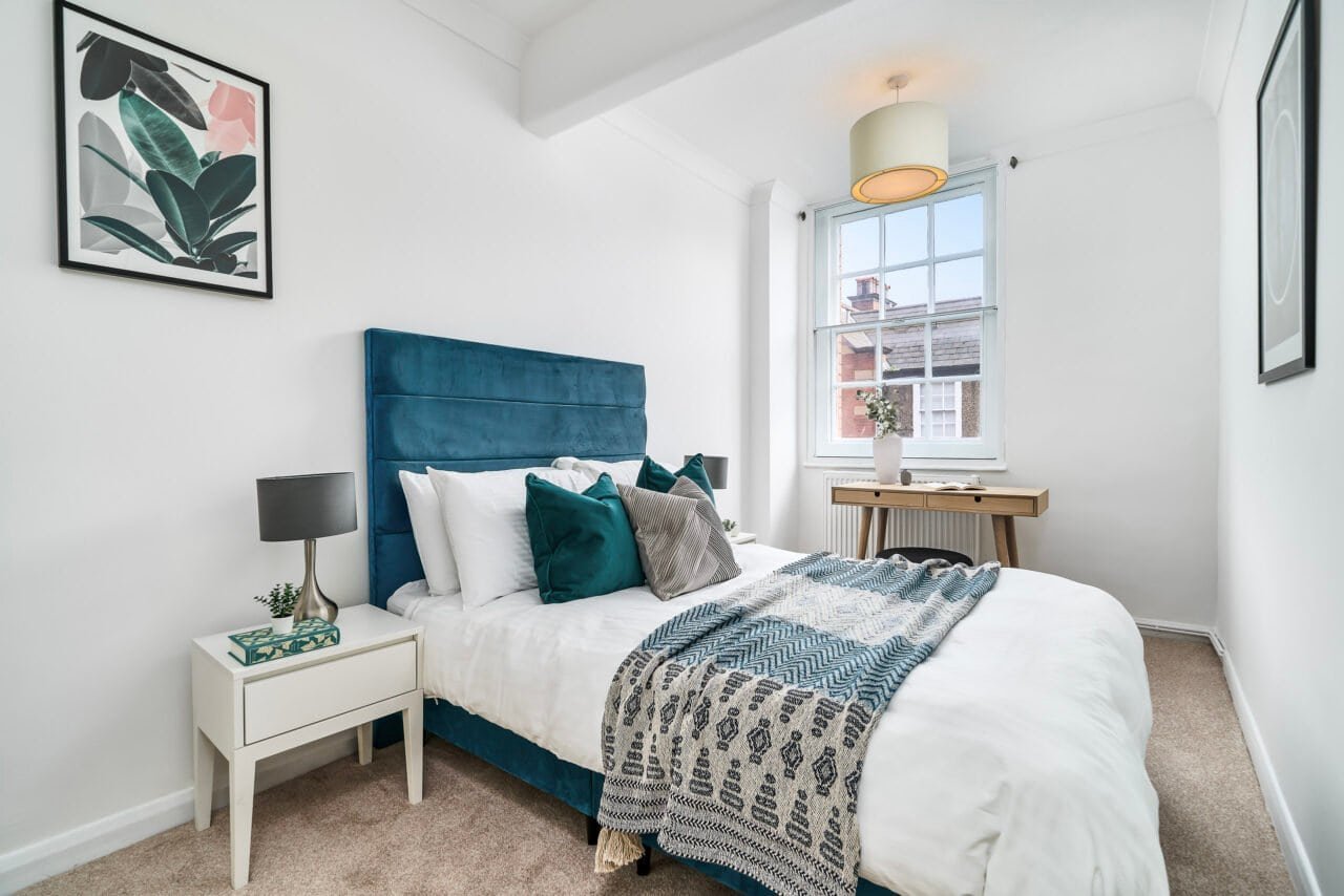 A comfortable bedroom with a blue upholstered headboard, white walls, and natural light.