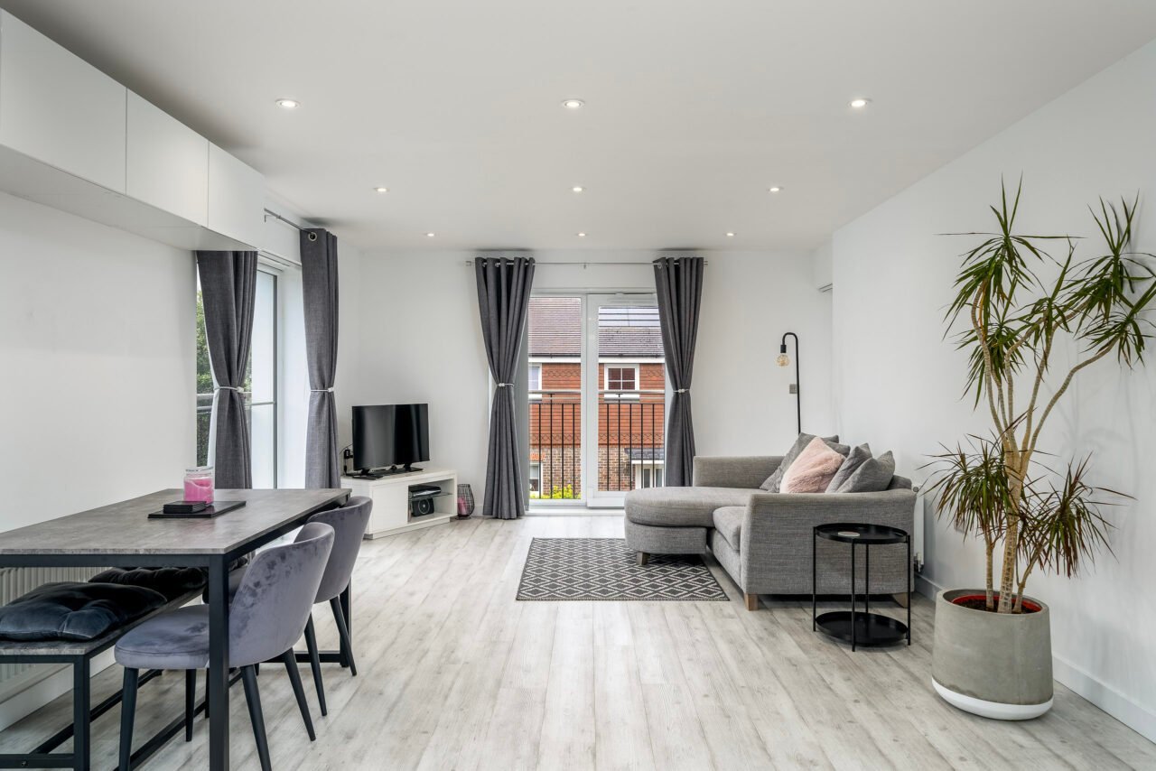 A modern, bright living room with grey sofa, dining area, and large windows with curtains.