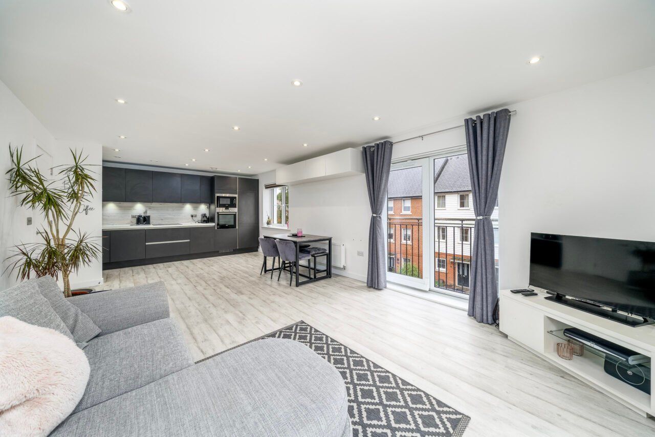 Modern open-plan living room with large windows, grey sofa, and flat-screen TV.