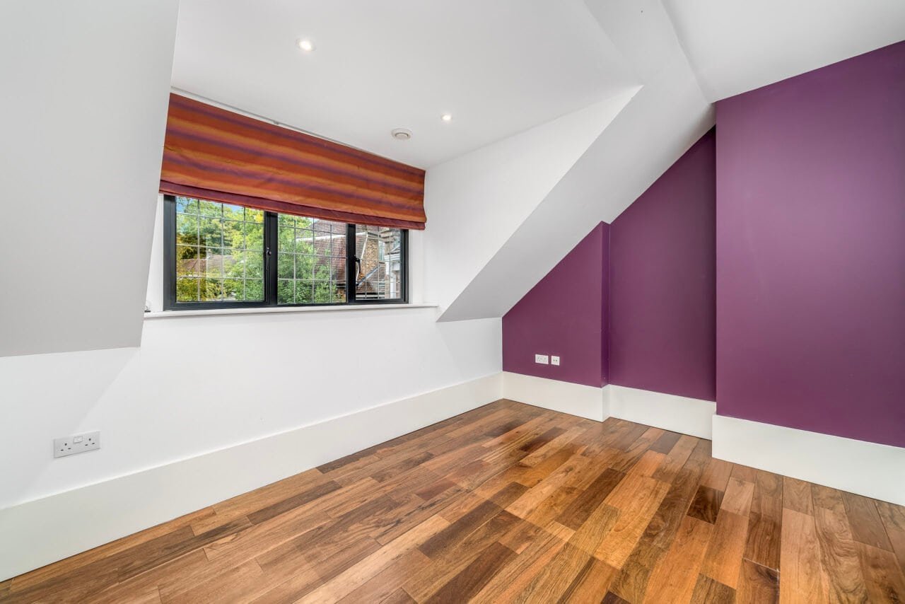 A spacious attic room with white walls, purple accent wall, wooden flooring, and window with blinds.