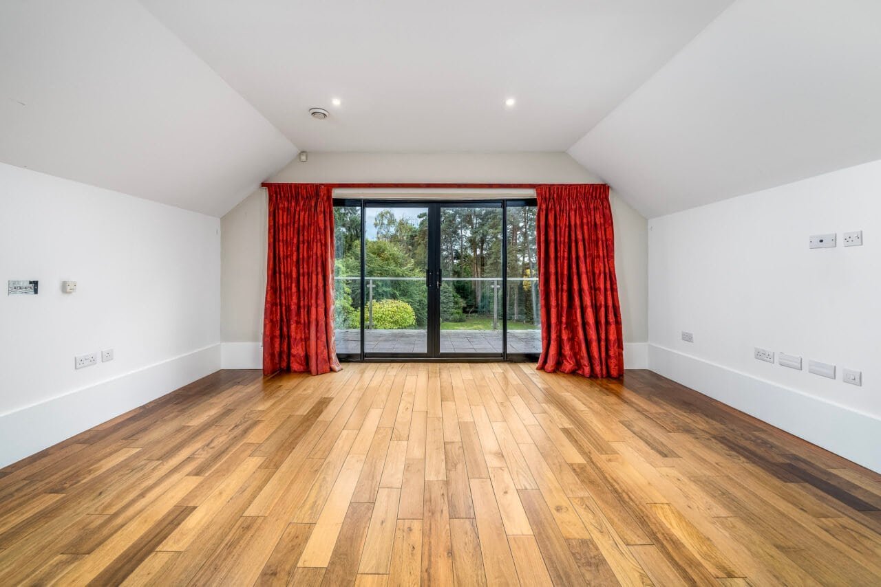 Spacious empty room with wooden flooring and large glass doors with red curtains.