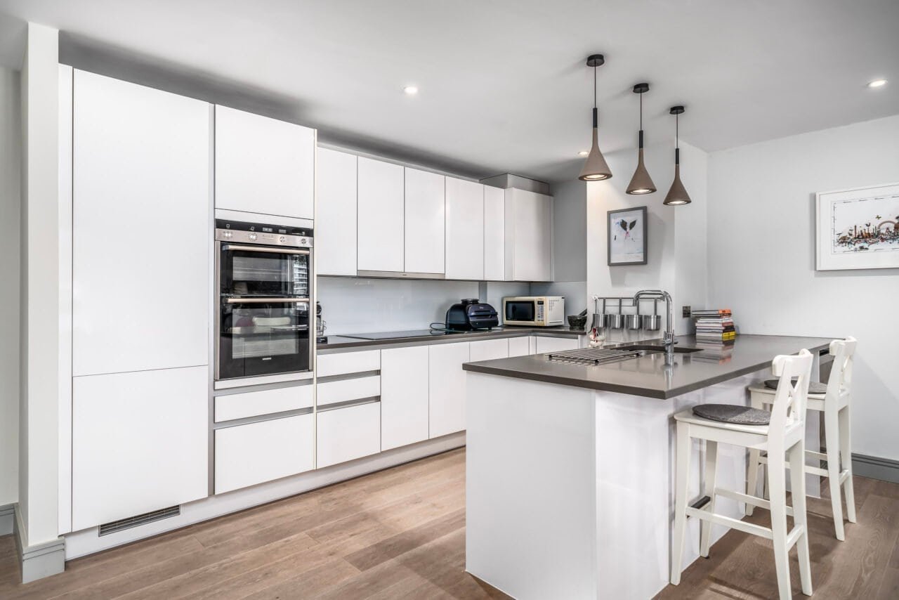 A modern white kitchen with built-in appliances and a central island for cooking and dining.