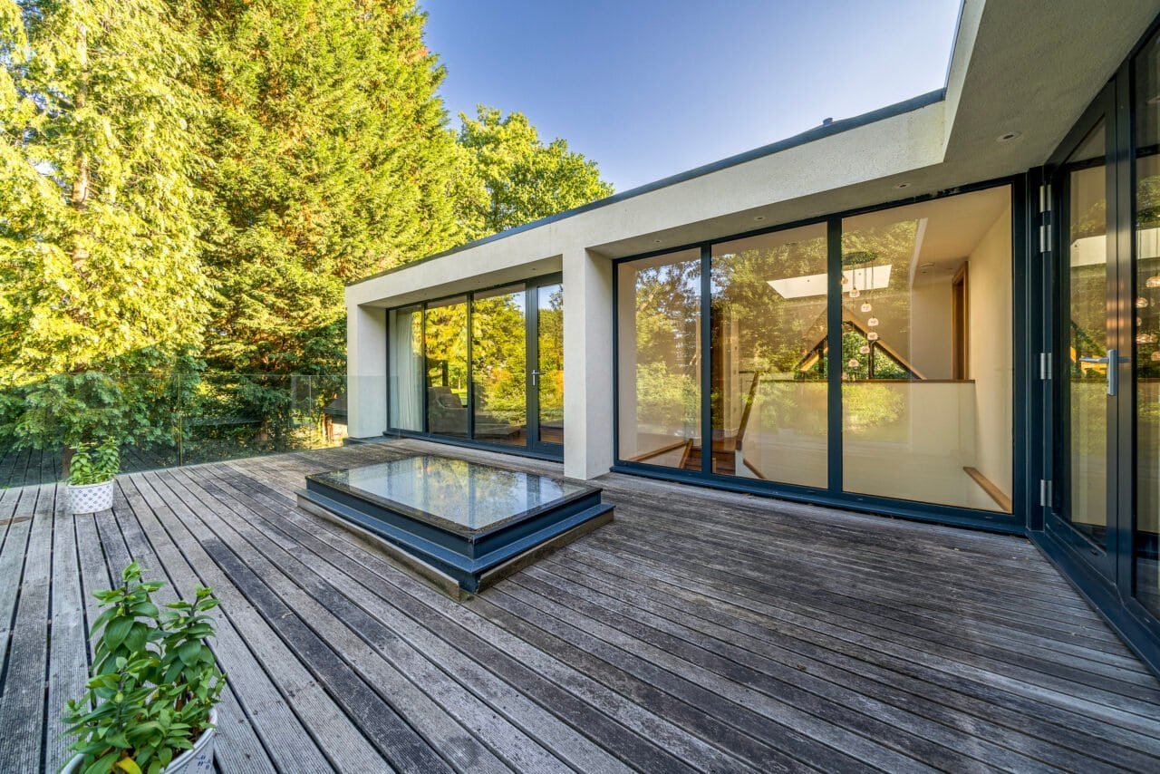 Modern house with large glass windows and wooden deck in a lush green garden.