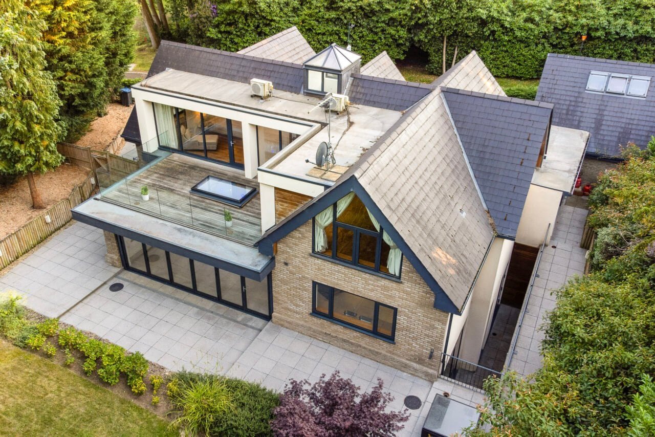Modern detached house with rooftop terrace and large glass windows, surrounded by greenery.