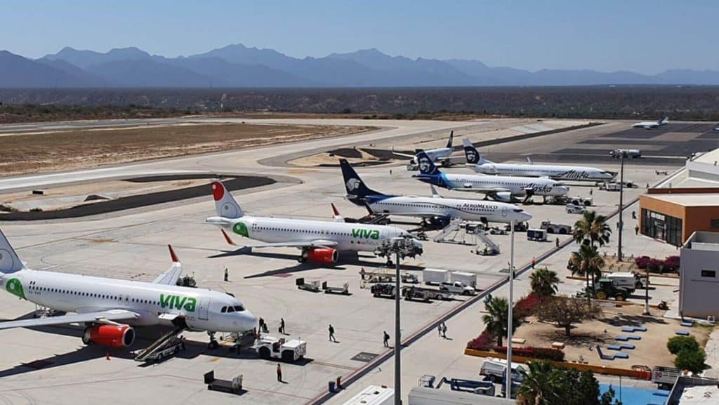 Aerial View Of The Los Cabos Airport Tarmac Showing Multiple Parked Commercial Jetliners From Airlines Including Viva Aerobus, Aeroméxico, And Alaska Airlines, With Rugged Mountains In The Background.