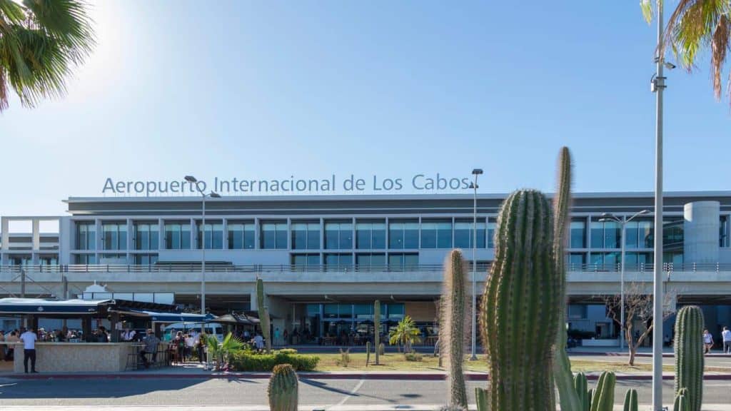 The Modern Facade Of Aeropuerto Internacional De Los Cabos (Los Cabos International Airport) Or The Cabos Mexico Airport Seen Past Large Desert Cacti And Palm Trees Under A Bright Blue Sky.